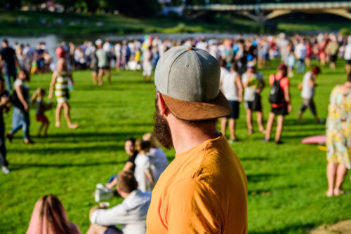 Hipster in cap happy celebrate event fest or festival. Summer fest. Man bearded hipster in front of crowd. Open air concert. Fan zone. Music festival. Entertainment concept. Visit summer festival.
