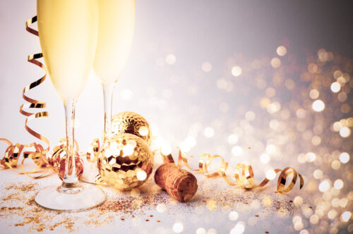 Two champagne flutes with golden christmas ornaments, streamer and lights book on a purple background